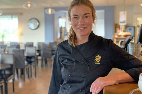 Annika Gripenberg står lutad mot receptionsdisken och ler mot kameran. I bakgrunden syns en tom restaurang.