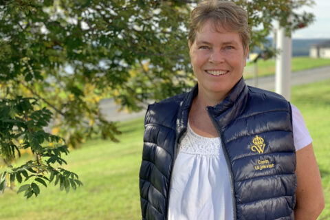 Carin Liljeqvist, klädd i marinblå väst och vit skjorta, står utomhus framför ett lövträd och ler mot kameran.