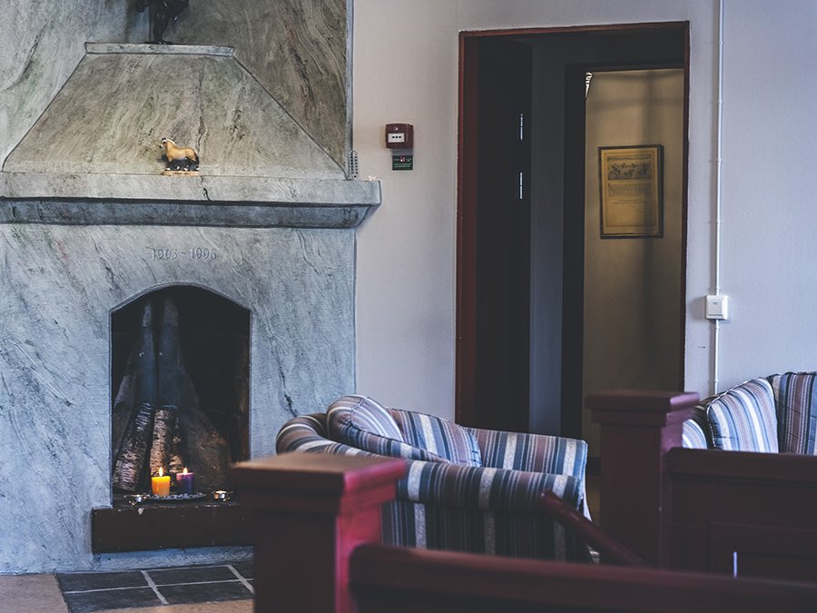 Täljstenskamin med tända ljus i. I förgrunden syns ett trappräcke i trä och en fotölj