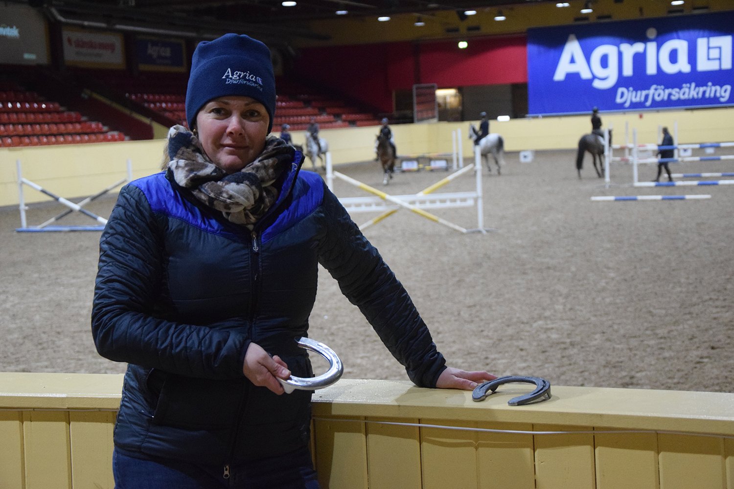 Hovslagare Jennie Sundin syns här i mössa och vinterkläder står på läktaren i ett ridhus och håller i en hästsko. I bakgrunden syns uppställda hinder och hästekipage.