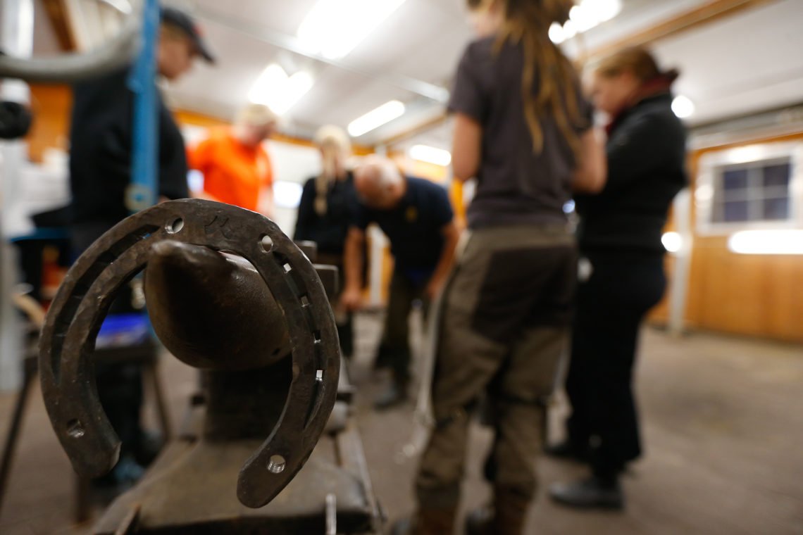 Städ med järnsko syns i förgrunden. I bakgrunden en hovslagarlärare omgiven av elever i skohallen