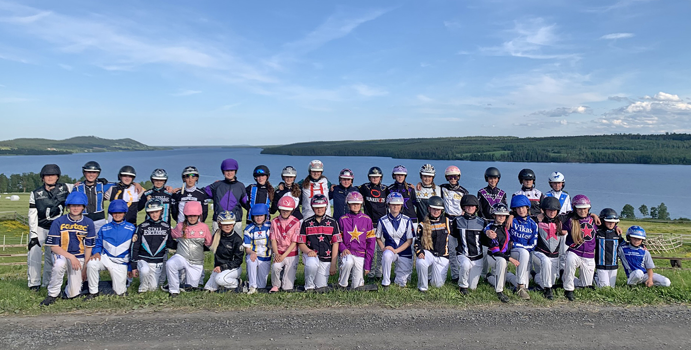 Gruppbild på lägerungdomar i kuskdressar och med hjälm på. I bakgrunden syns en sjö