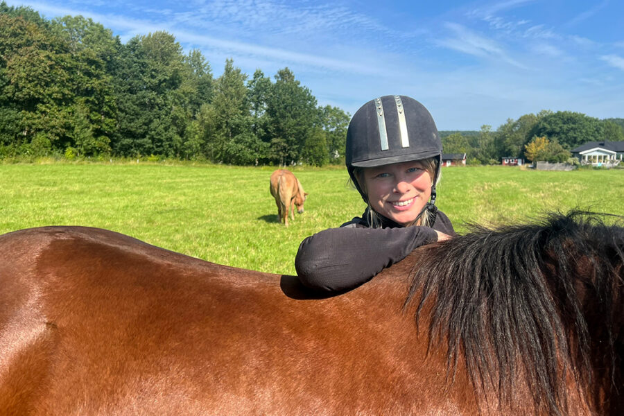 En person i ridhjälm ler medan hon lutar sig mot en brun häst på ett gräsbevuxet fält, med en annan häst som betar under en blå himmel.