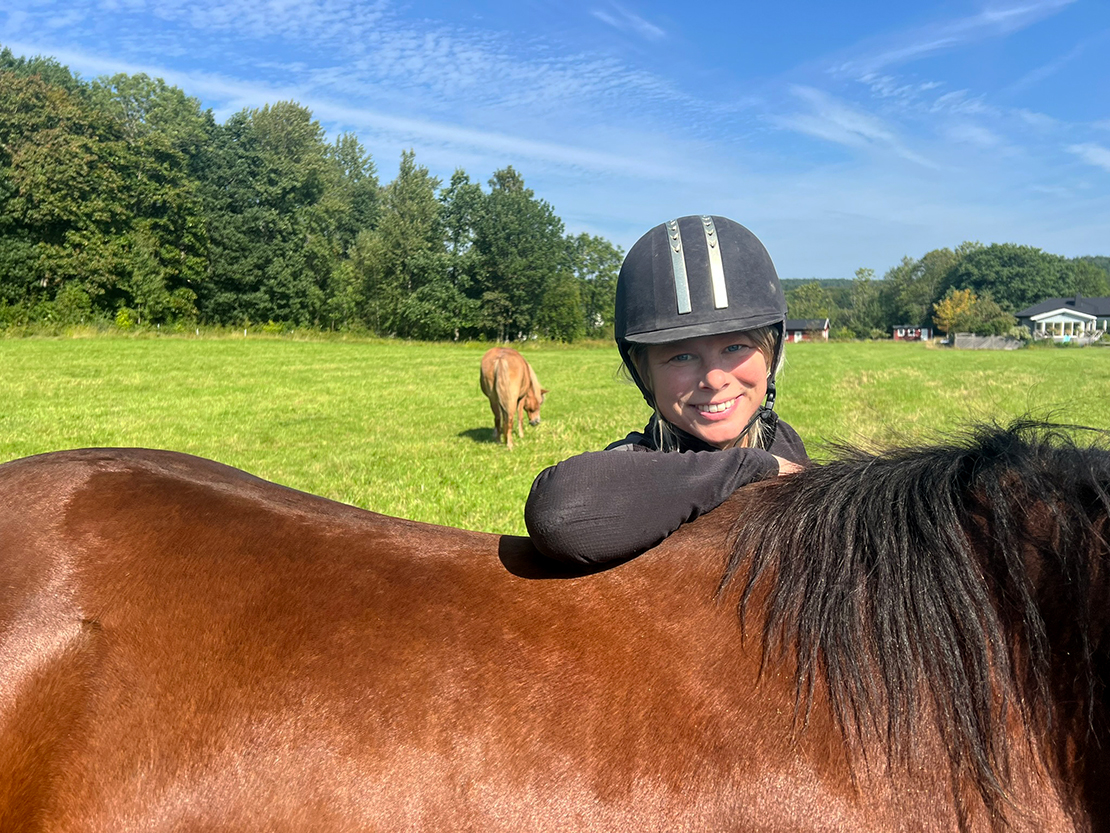 En person i ridhjälm ler medan hon lutar sig mot en brun häst på ett gräsbevuxet fält, med en annan häst som betar under en blå himmel.