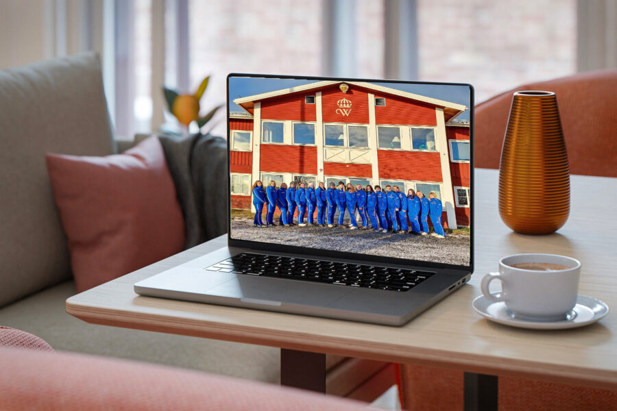 En laptop på ett bord visar ett gruppfoto av gymnasieelever i blå uniformer framför en röd byggnad; en kopp kaffe och en orange vas står bredvid laptopen.