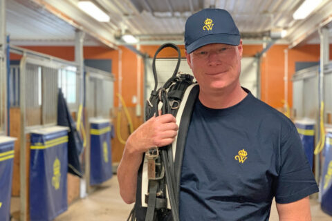 Man står i stallgång med en travsele över axeln. Han har blå keps och blå t-shirt med Wångens emblem på
