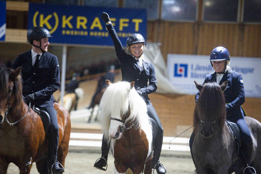 Tre tävlingsklädda ryttare på islandshästar i ridhuset under prisutdelning; den mittersta ryttaren höjer en arm, medan de andra tittar på och ler under Wångens islandshästdagar.