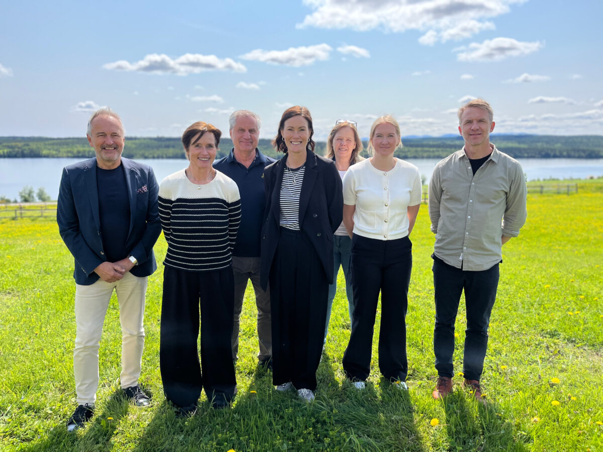 En grupp personer står utomhus en solig dag på en gräsmatta. I bakgrunden syns hästhagar och en sjö.