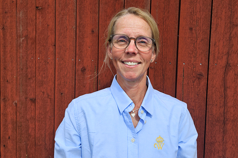 Leende kvinna med glasögon står mot en röd vägg utomhus. Hon har en ljusblå skjorta med Wångens emblem på.