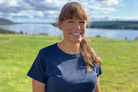 Kvinna med mörkt långt hår i tofs och lugg ler mot kameran en solig dag utomhus på Wången. Hon har på sig en blå t-shirt med Wångens sigill. I bakgrunden syns sjön.