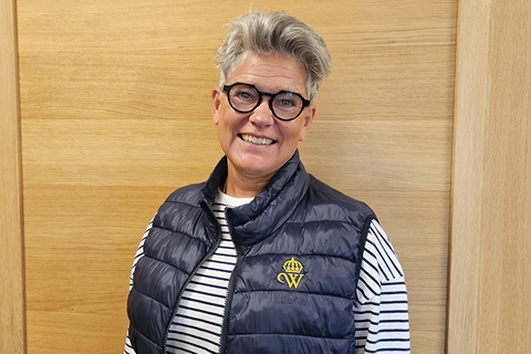 Kvinna med kort hår och glasögon står mot en vägg inomhus. Hon har blå väst med Wångens emblem på.