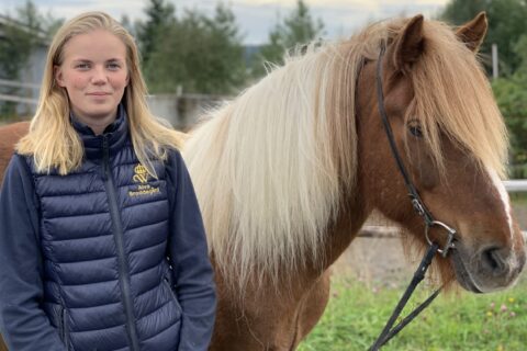 Ung kvinna i väst med Wångens sigill står utomhus och håller i tyglarna till en brunskäckig islandshäst