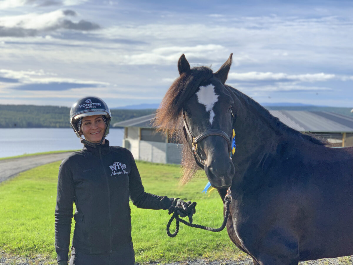 Kvinna i svart jacka och hjälm står bredvid en svart kallblodstravare utomhus en solig dag. I bakgrunden syns en sjö och en skrittmaskin.