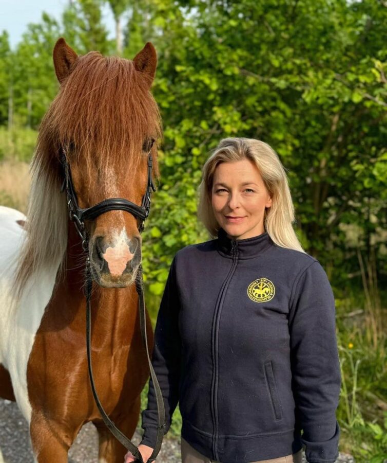 Blond kvinna i blå tröja med Svenska Islandshästförbundets logtoyp på står bredvid en brunskäckig islandshäst