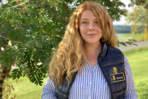 Tina Larsson, en kvinna med långt rött hår, klädd i randig skjorta och mörk väst med broderad badge, står utomhus framför gröna bladverk.