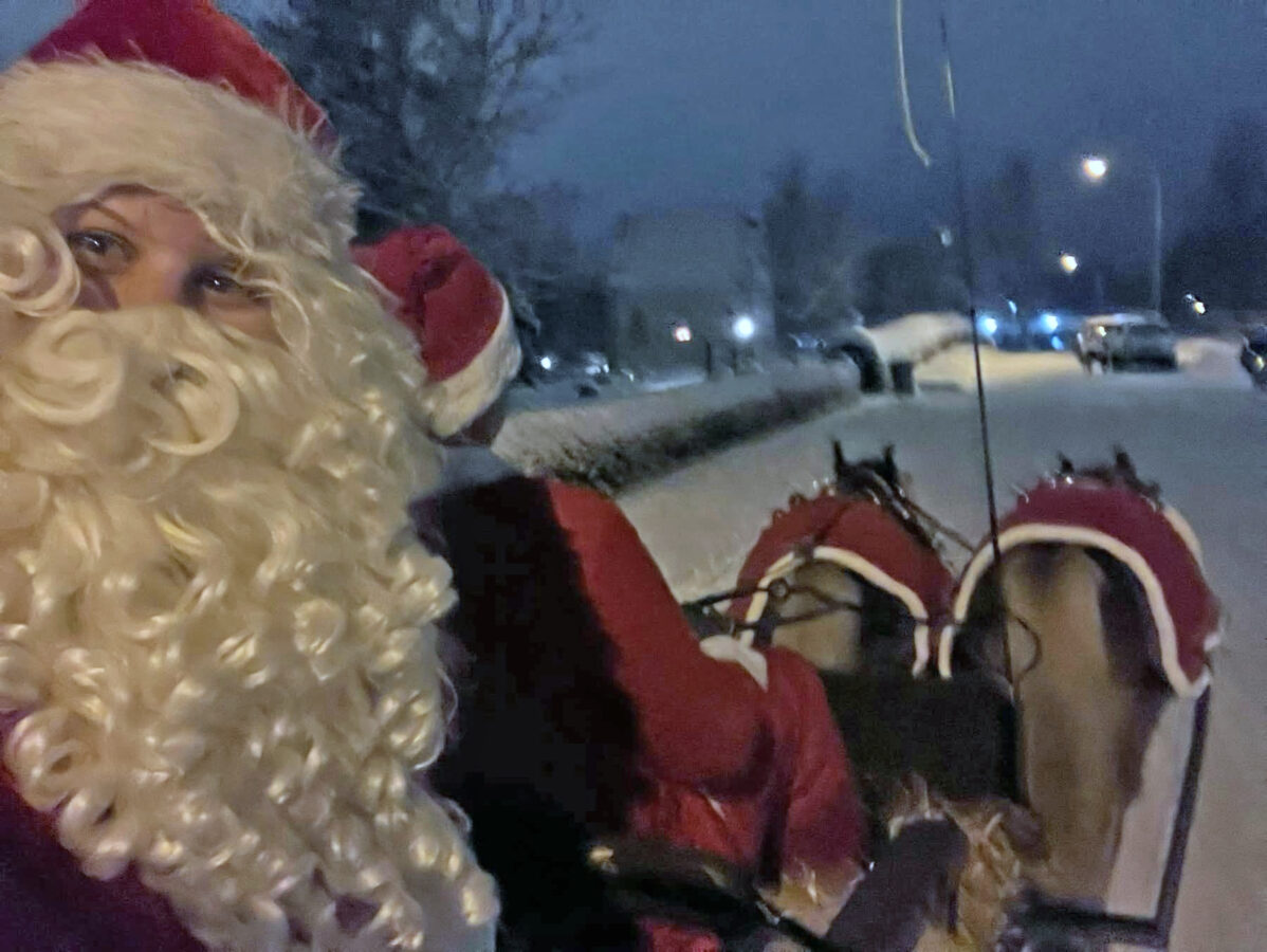 Person klädd som jultomten sitter i en Tylla 505 hästdragen släde på en snöig gata på natten, med två hästar i röda täcken synliga framför.