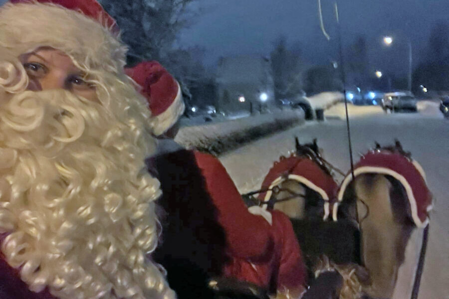 Person klädd som jultomten sitter i en Tylla 505 hästdragen släde på en snöig gata på natten, med två hästar i röda täcken synliga framför.