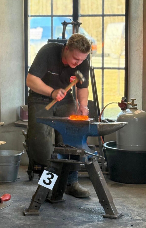 Man står vid ett städ med hammare i handen och en glödande järnsko ligger på städet