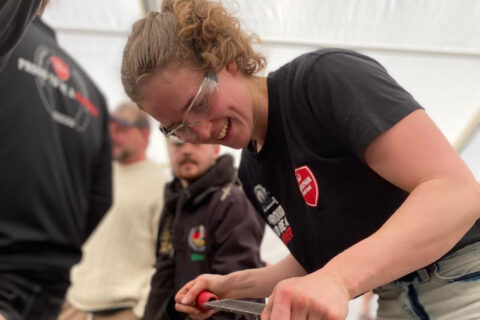 En person med skyddsglasögon och svart skjorta använder ett verktyg, medan andra personer syns i bakgrunden inne på ett tältområde.