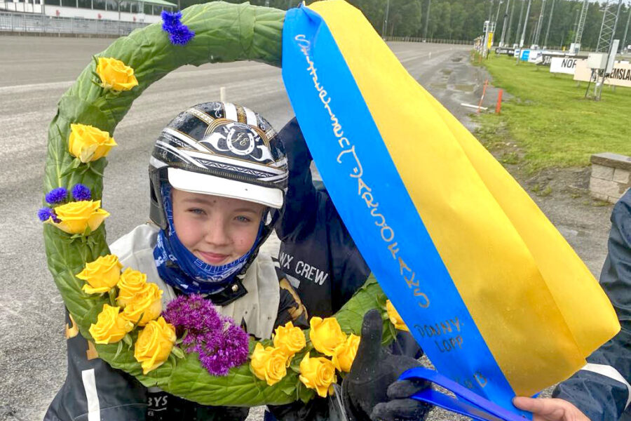 En ung travkusk med hjälm och tävlingskläder håller i en stor blommig segerkrans och ett blågult band.