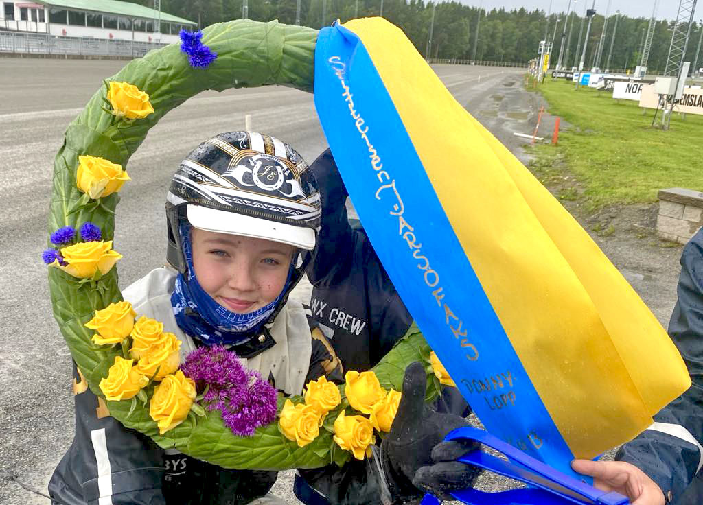 En ung travkusk med hjälm och tävlingskläder håller i en stor blommig segerkrans och ett blågult band.
