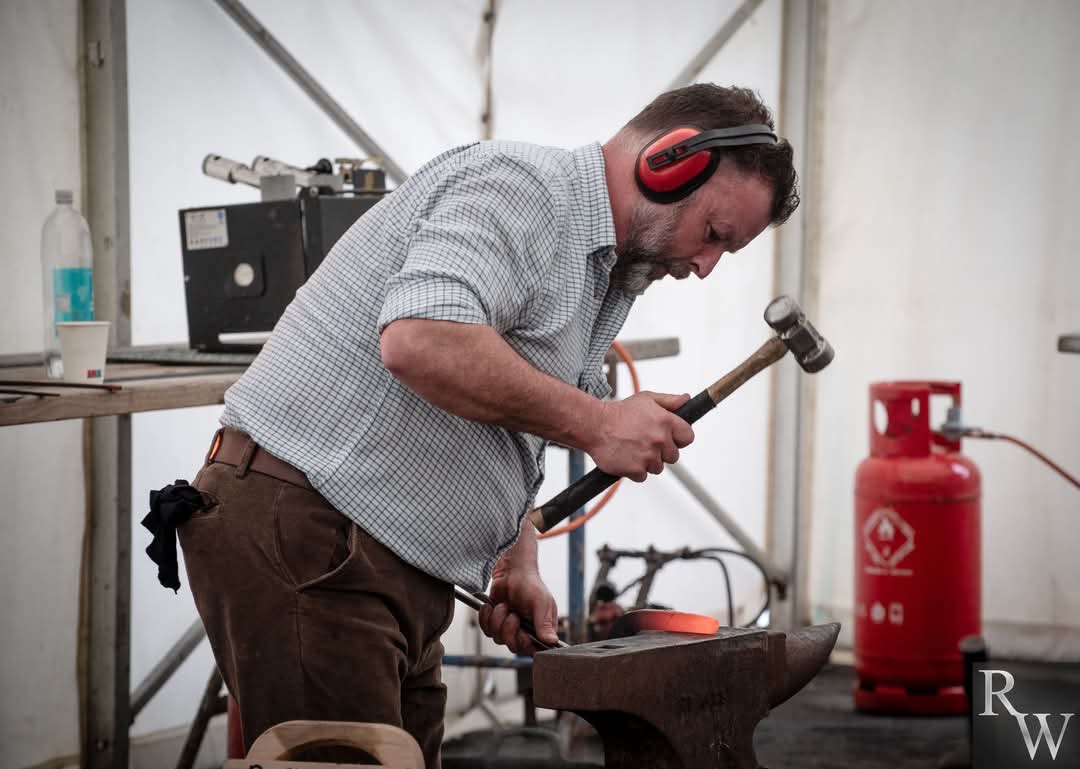 En man med hörselkåpor använder en hammare för att forma het metall på ett städ. en röd gasflaska syns i bakgrunden.