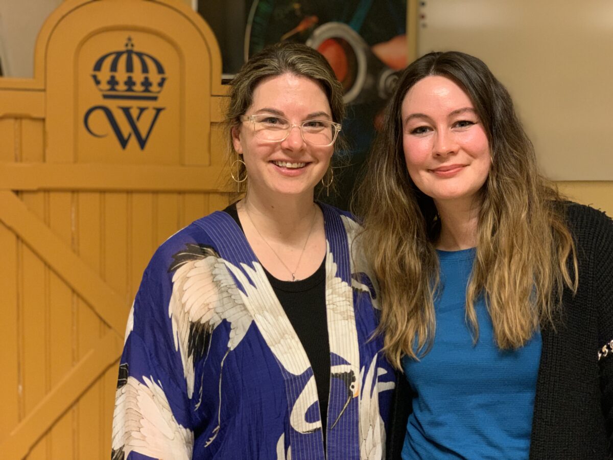 Två kvinnor står sida vid sida och ler mot kameran, bakom syns en gultalarstol med Wångenemblemet på