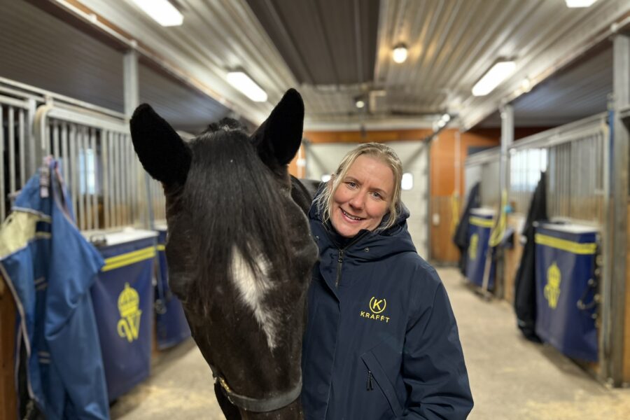 Kvinna i blå jacka med KRAFFTs logotyp på står i stallgången på Wången med en mörkbrun varmblodstravare