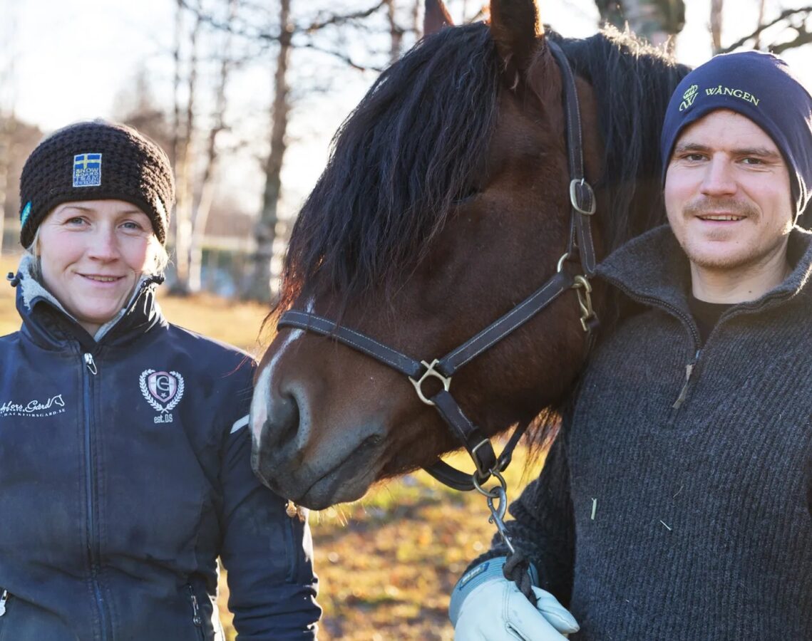 En kvinna och en man ler och tittar in i kameran. De står utomhus med en nordsvensk brukshäst emellan sig