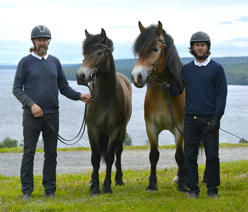 Två män i hjälm och tröja står utomhus på Wången och håller i tyglarna på två hästar, med Alsensjön och skog i bakgrunden.