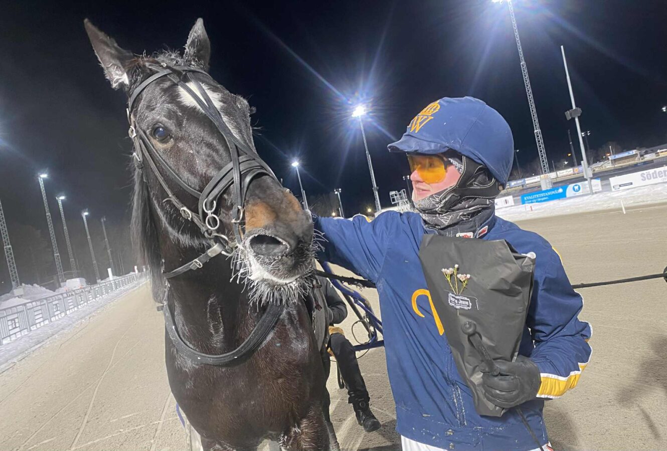 En travkusk i en blå dress med Wångens gula emblem på står bredvid Villiam Avenue på en travbana på natten och håller i en blombukett.