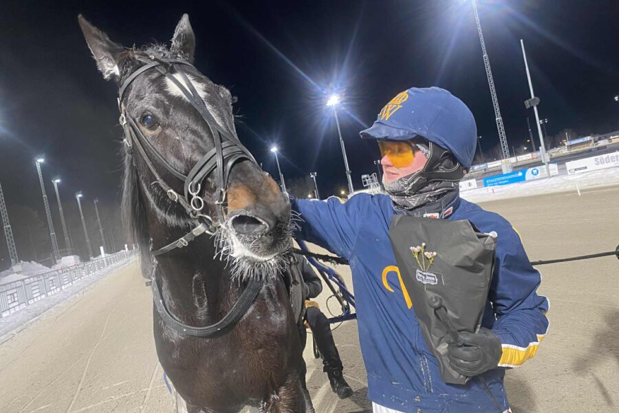 En travkusk i en blå dress med Wångens gula emblem på står bredvid Villiam Avenue på en travbana på natten och håller i en blombukett.