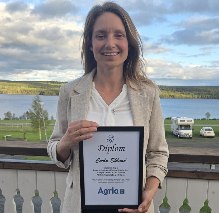 En kvinna står utomhus, ler och håller i ett inramat stipendiatdiplom med Agrias logotyp på. I bakgrunden syns en sjö, ett fält och en husbil under en molnig himmel.