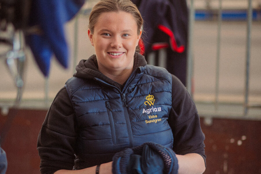 Ebba, årets Wångenambassadör, med brunt hår, bär blå väst med Wångens sigill och Agrias logotyp på, står i en stallgång och ler mot kameran.