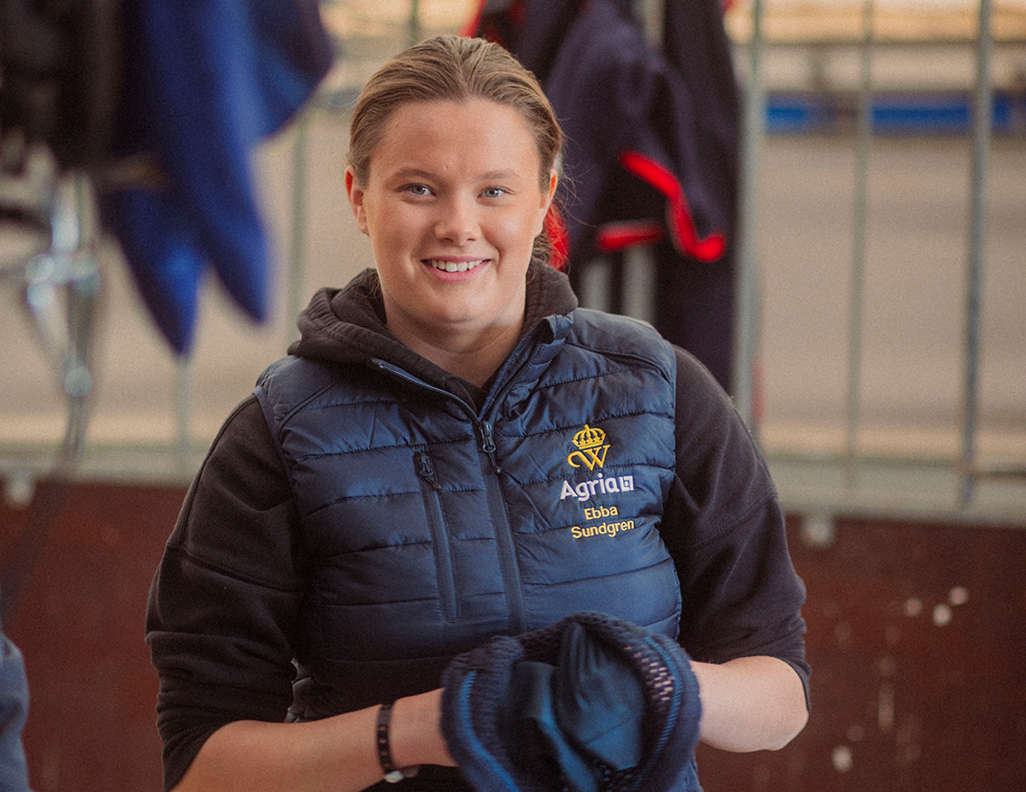 Ebba, årets Wångenambassadör, med brunt hår, bär blå väst med Wångens sigill och Agrias logotyp på, står i en stallgång och ler mot kameran.