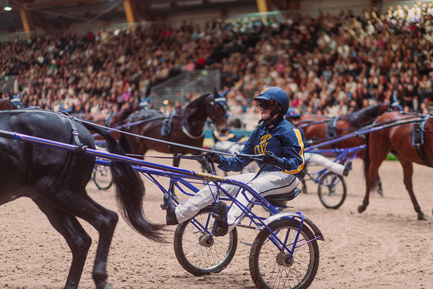 Ebba iförd hjälm och Wångens kuskdress, kör en sulky dragen av en häst i en inomhusarena med en stor publik i bakgrunden.