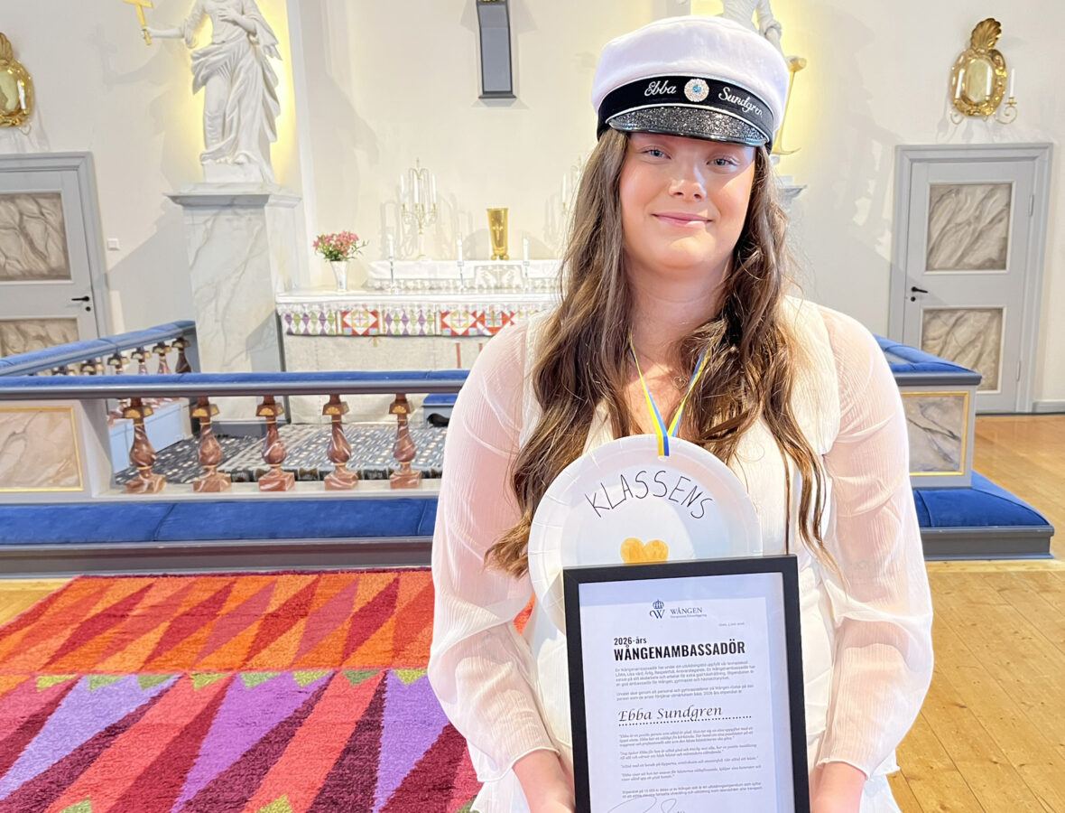En ung kvinna med brunt långt hår och studentmössa och vita kläder står i en kyrka. Runt halsen har hon en papptallrik där det står Klassens guldhjärta på och i handen håller hon ett inramat diplom för Wångenambassadören