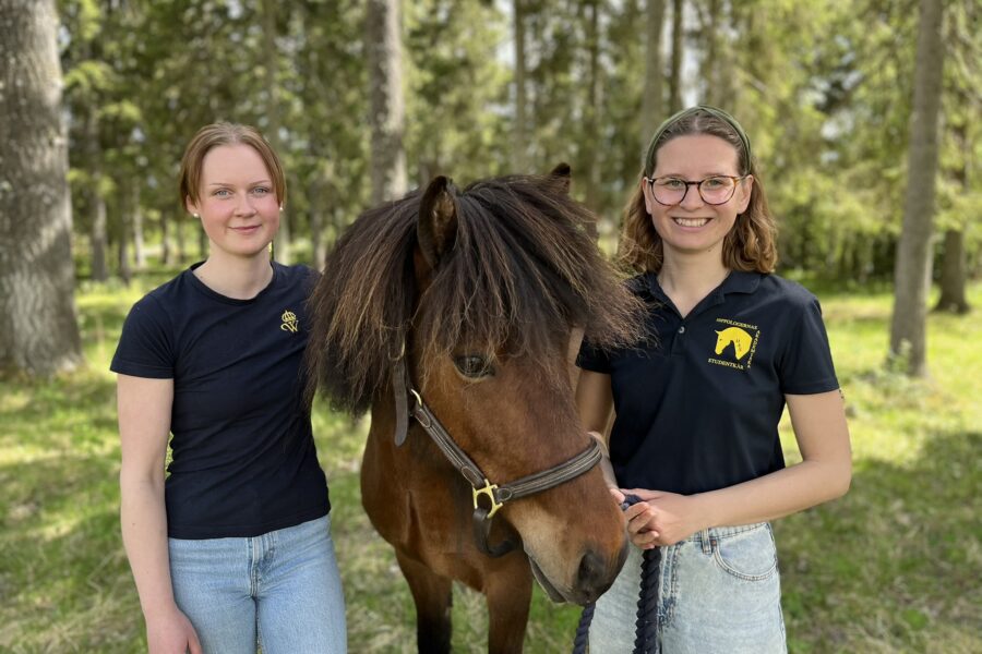Två unga kvinnor står på var sin sida om en brun islandshäst. De står i en skog en solig dag och ler mot kameran. De nyexaminerade hippologerna har genomfört en studie om musik påverkar rytmen i tölten.