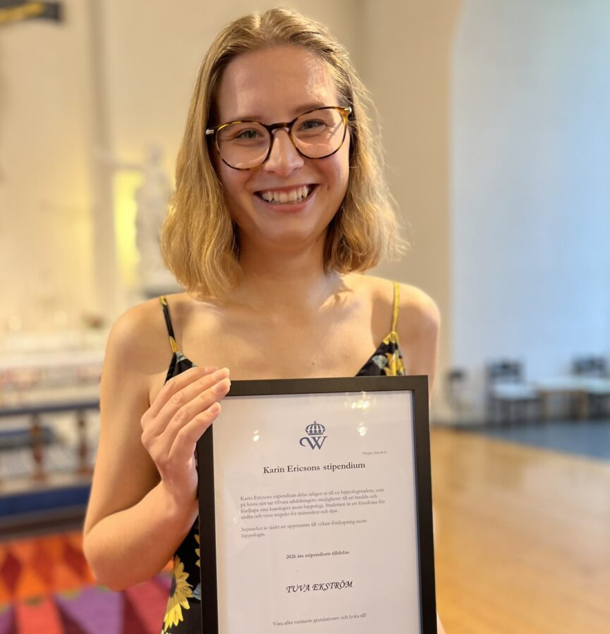 Ung kvinna i page och glasögon ler mot kameran. Hon står i Alsens kyrka och håller i ett inramat diplom. Hon har en klänning med solrosor på svart botten på sig. 