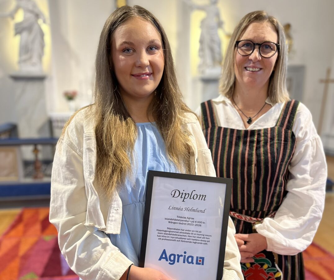 En ung kvinna i ljusblå klänning står i en kyrka. I handen håller hon ett inramat diplom från Agria. Bredvid henne står lärare Camilla Hed iklädd folkdräkt.
