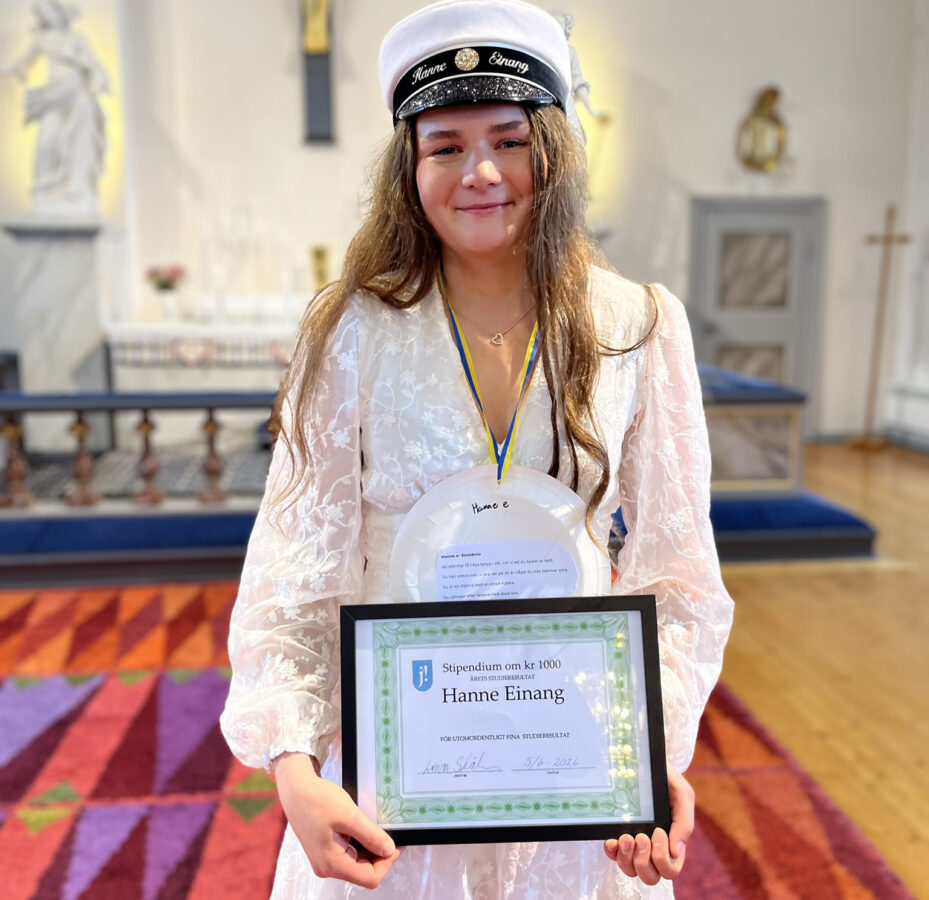 Ung kvinna med långt brunt hår, studentmössa och vit klänning står i Alsens kyrka och håller i ett inramat diplom med svart och grön ram.