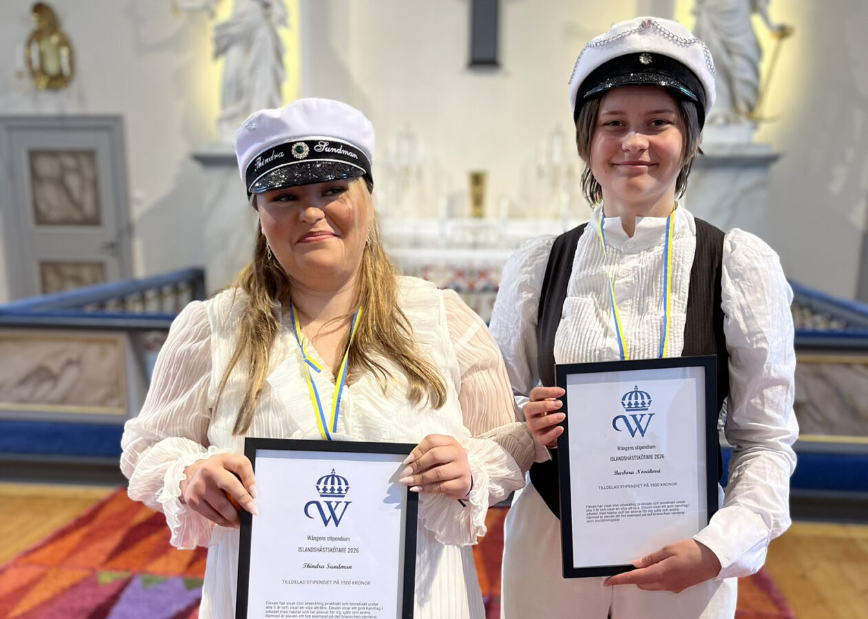 Två unga kvinnor står bredvid varandra i en kyrka och håller i varsitt inramat diplom med Wångens blå sigill på. 
