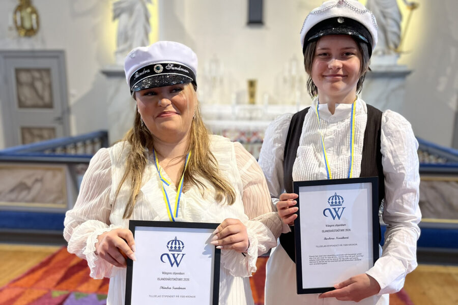 Två unga kvinnor står bredvid varandra i en kyrka och håller i varsitt inramat diplom med Wångens blå sigill på.