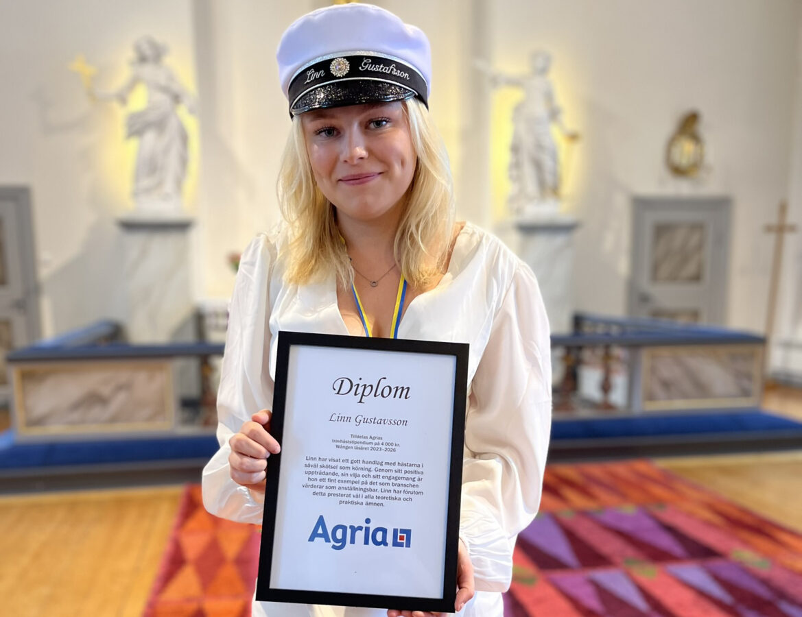 En ung blond kvinna med stundentmössa står i en kyrka. I handen håller hon ett diplom med Agrias logotyp på.