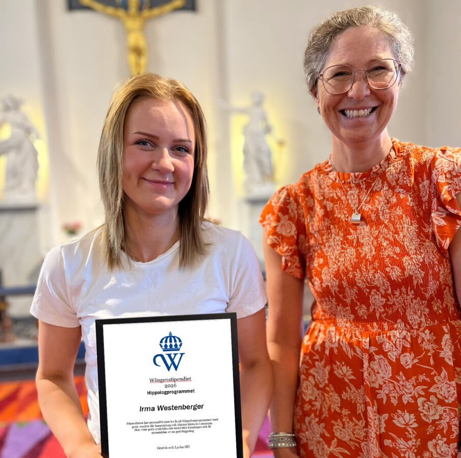 En ung kvinna i vit t-shirt håller i ett inramat diplom. Bredvid henne står lärare Joanna Sätter i orange mönstrad klänning. De ler mot kameran stående i Alsens kyrka. 