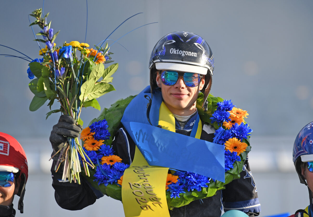 Kille i hjälm och kuskdress och spegelsolglasögon håller upp en blomsterbukett och har en segerkrans runt halsen