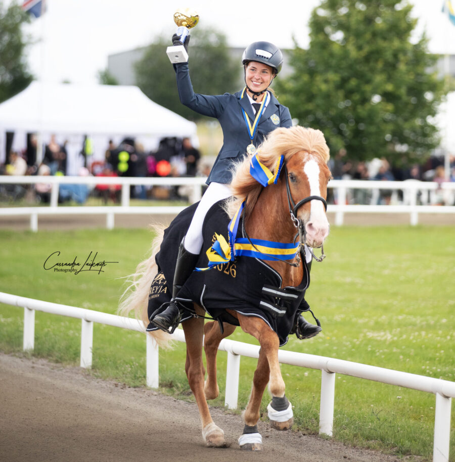 Ung kvinna i kavaj och vita ridbyxor rider en isabellfärgad islandshäst . Hästen har ett vinnartäcke och Saga håller upp en pokal i luften
