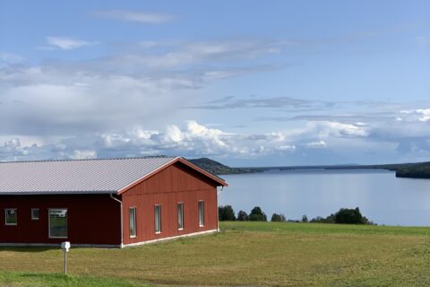 Röd byggnad på en gräskulle med utsikt över en sjö under blå himmel med moln