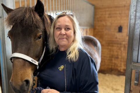 Keena står i stallet i mörkblå Wångentröja hon håller i islandshästen Prins som har en reflexgrimma från Agria på sig