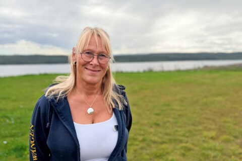 Leende kvinna med glasögon står på en gräsyta vid en sjö under molnig himmel